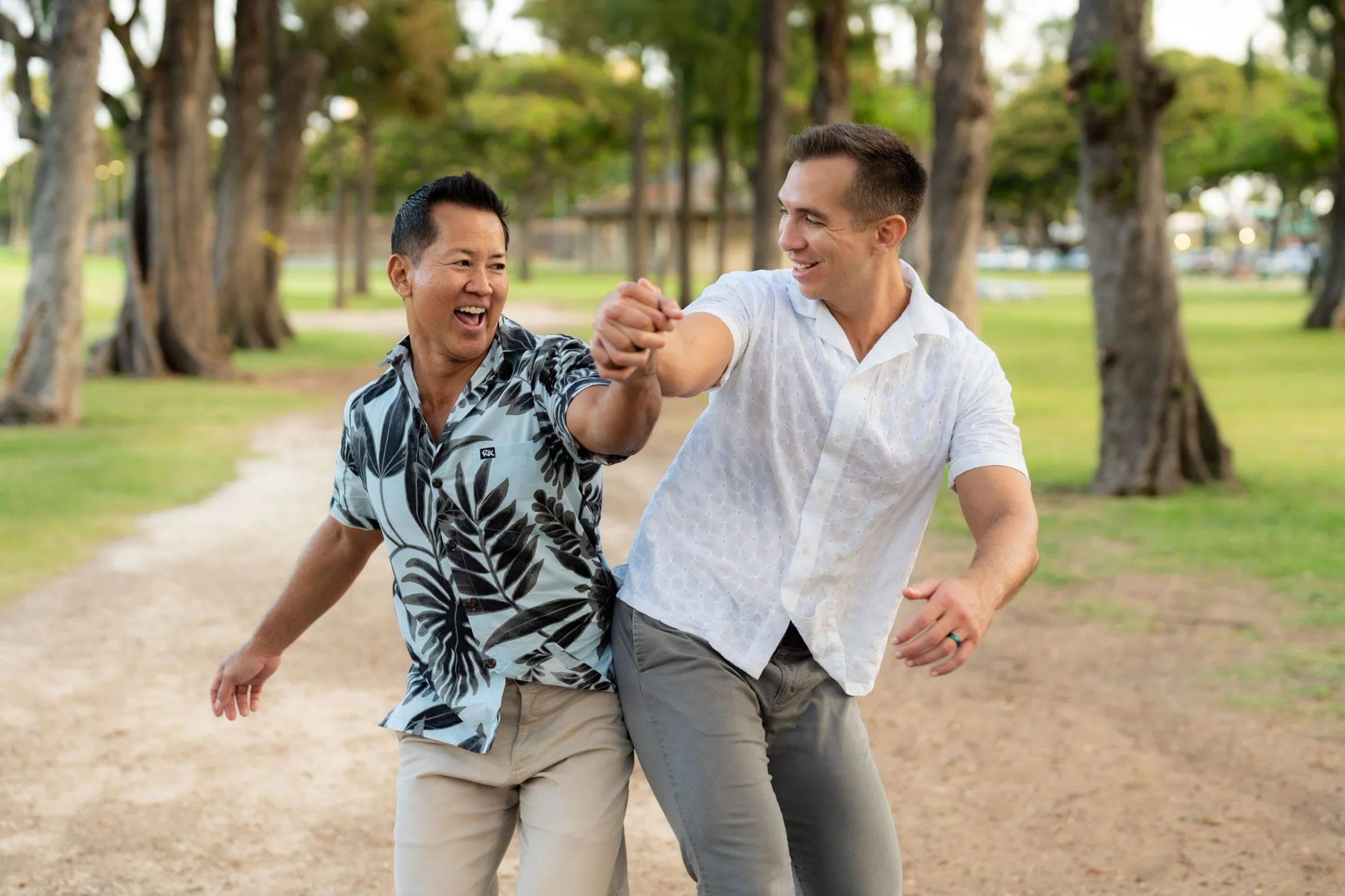 Two men laugh as they dance together under the trees in a park.