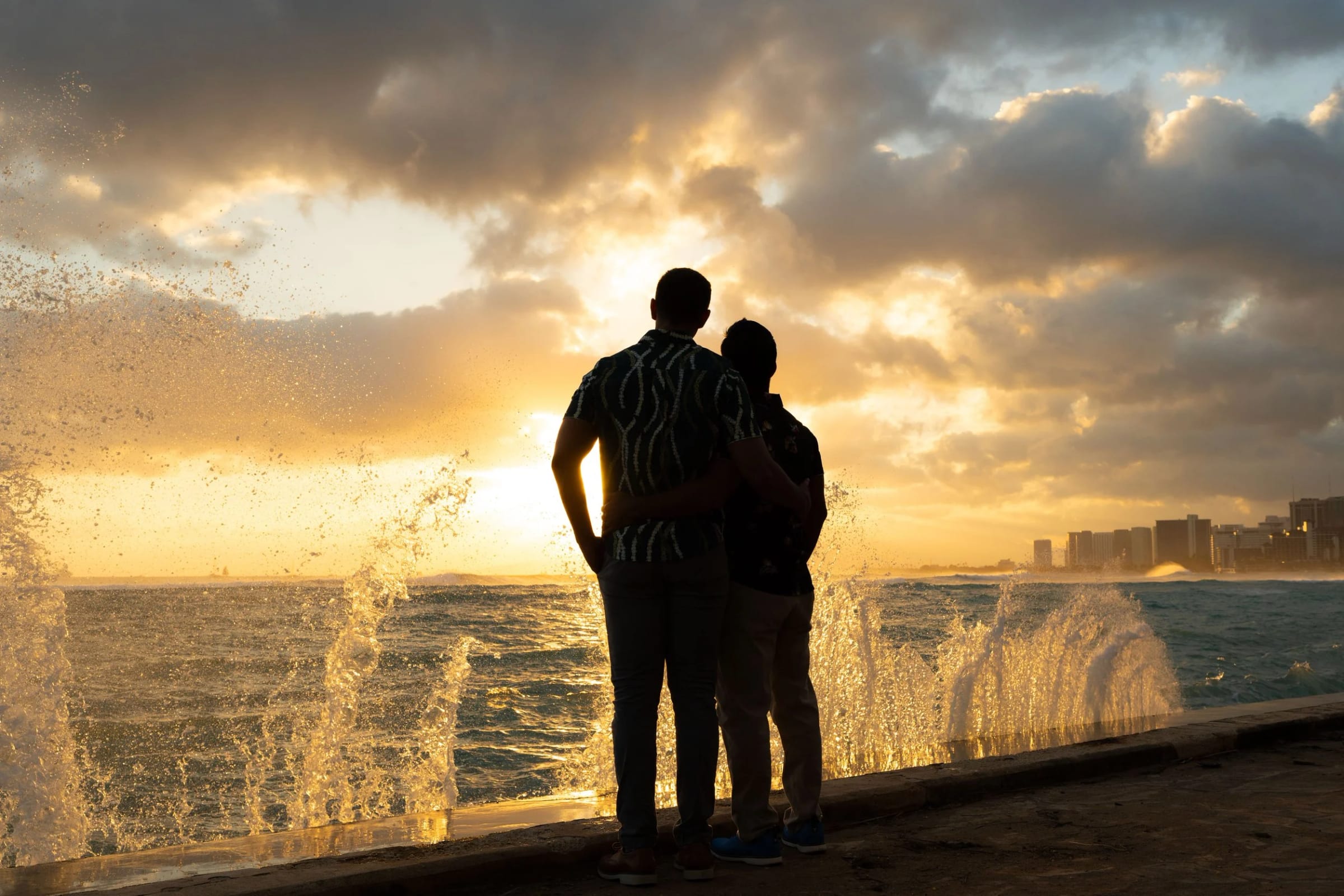 A couple in silhouette on a seawall at sunset as a wave breaks beside them.